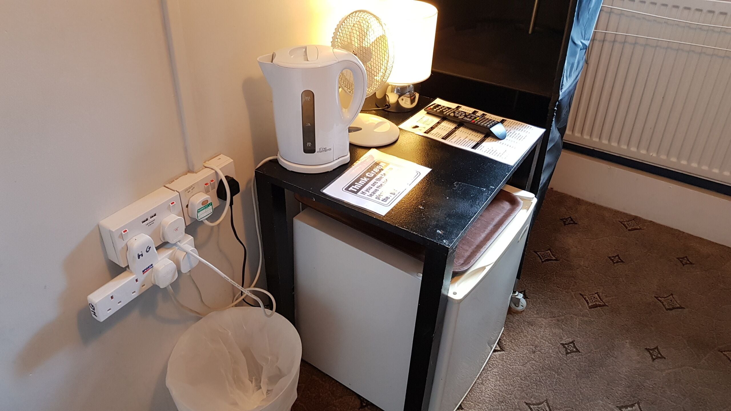 Eine kleine Küchenzeile mit Wasserkocher, Ventilator und Mikrowelle auf einer schwarzen Arbeitsplatte. Ein Mülleimer und eine Steckdose mit mehreren Steckplätzen sind vorhanden.
