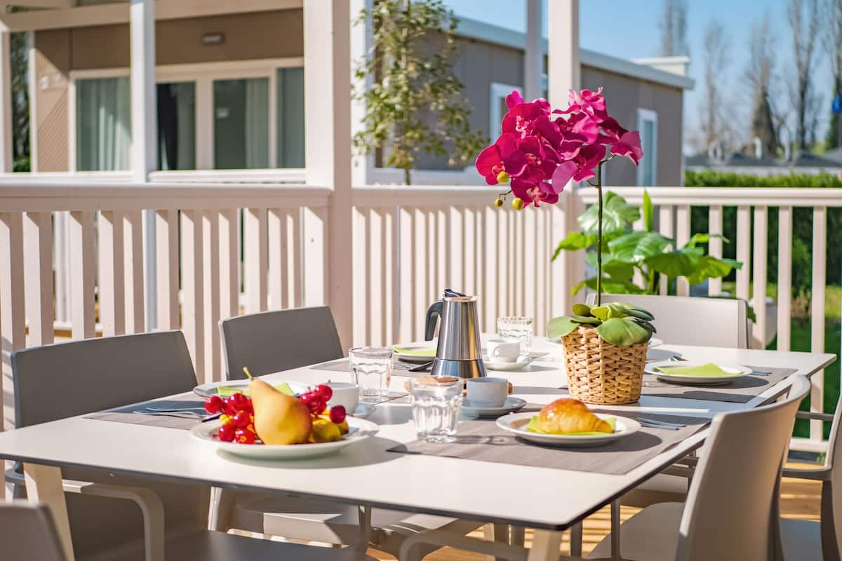 Mesa de comedor al aire libre con mantel blanco, cafetera, platos con comida y planta en maceta con flores rosas.