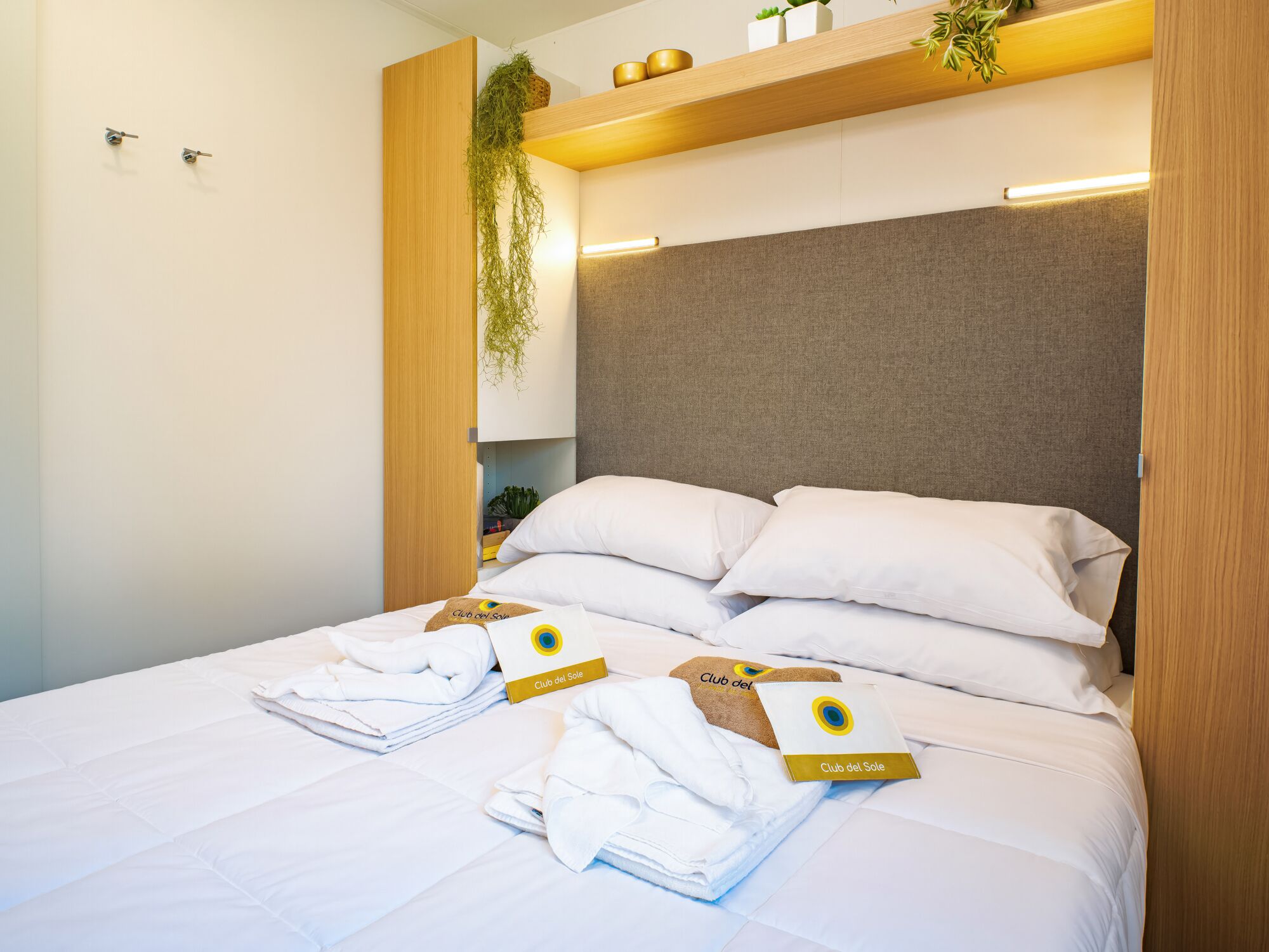 Una cama bien hecha con ropa de cama blanca y almohadas, cabecera de madera y estante con plantas y adornos.