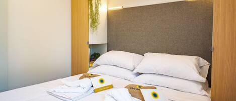 Una cama bien hecha con ropa de cama blanca y almohadas, cabecera de madera y estante con plantas y adornos.