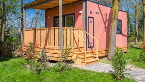 Une petite cabane à un seul étage, dotée d’une terrasse en bois et d’un porche couvert, entourée d’arbres et de verdure.