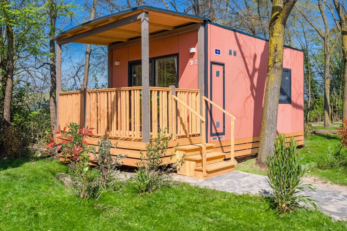 Una cabaña pequeña de una sola planta, con terraza de madera y porche cubierto, rodeada de árboles y vegetación.