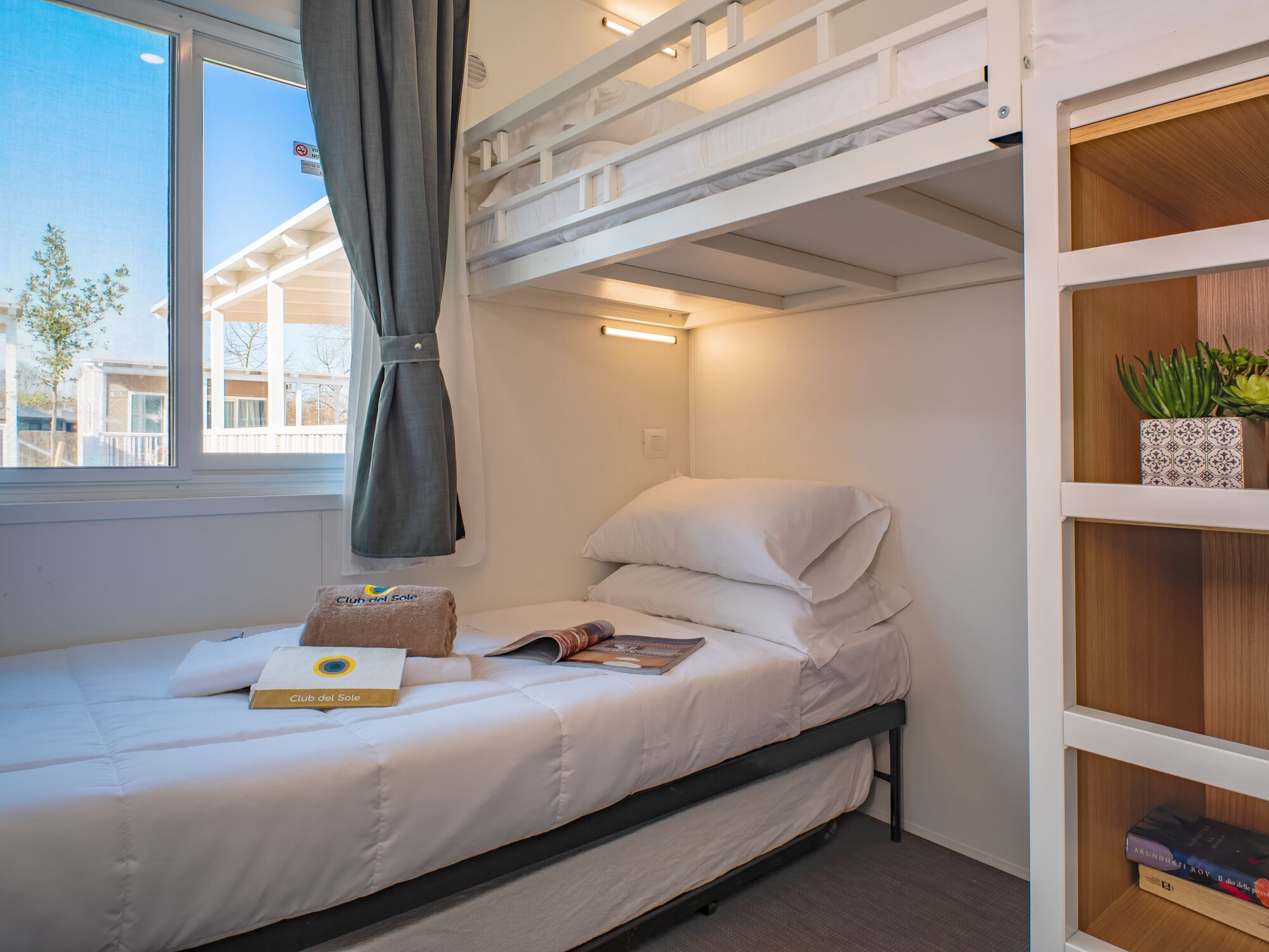 Ein Einzelbett mit weißer Matratze und Kissen, ein Nachttisch mit Buch und Handtuch, ein Fenster mit Vorhang und eine Holzablage mit Pflanze und Büchern.