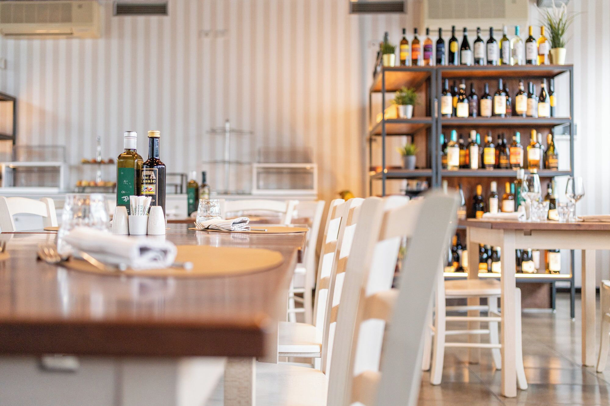 Un interior de restaurante con una larga mesa de comedor, sillas blancas y una zona de bar con estantes llenos de botellas.