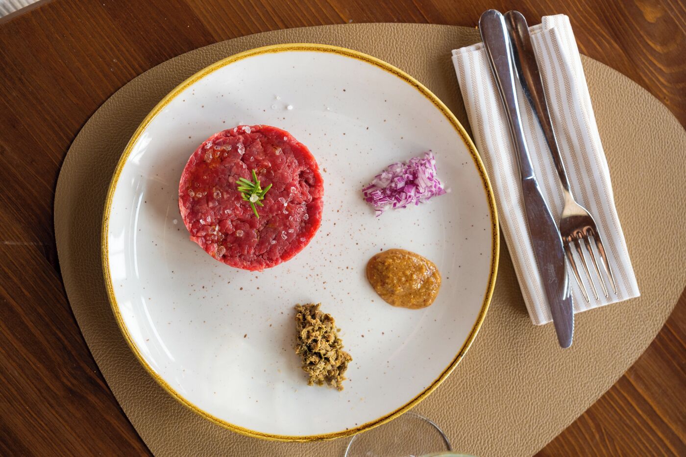 A plate with a red meat dish, a small pile of red onion, and a brown sauce, accompanied by a fork and knife on a beige napkin.