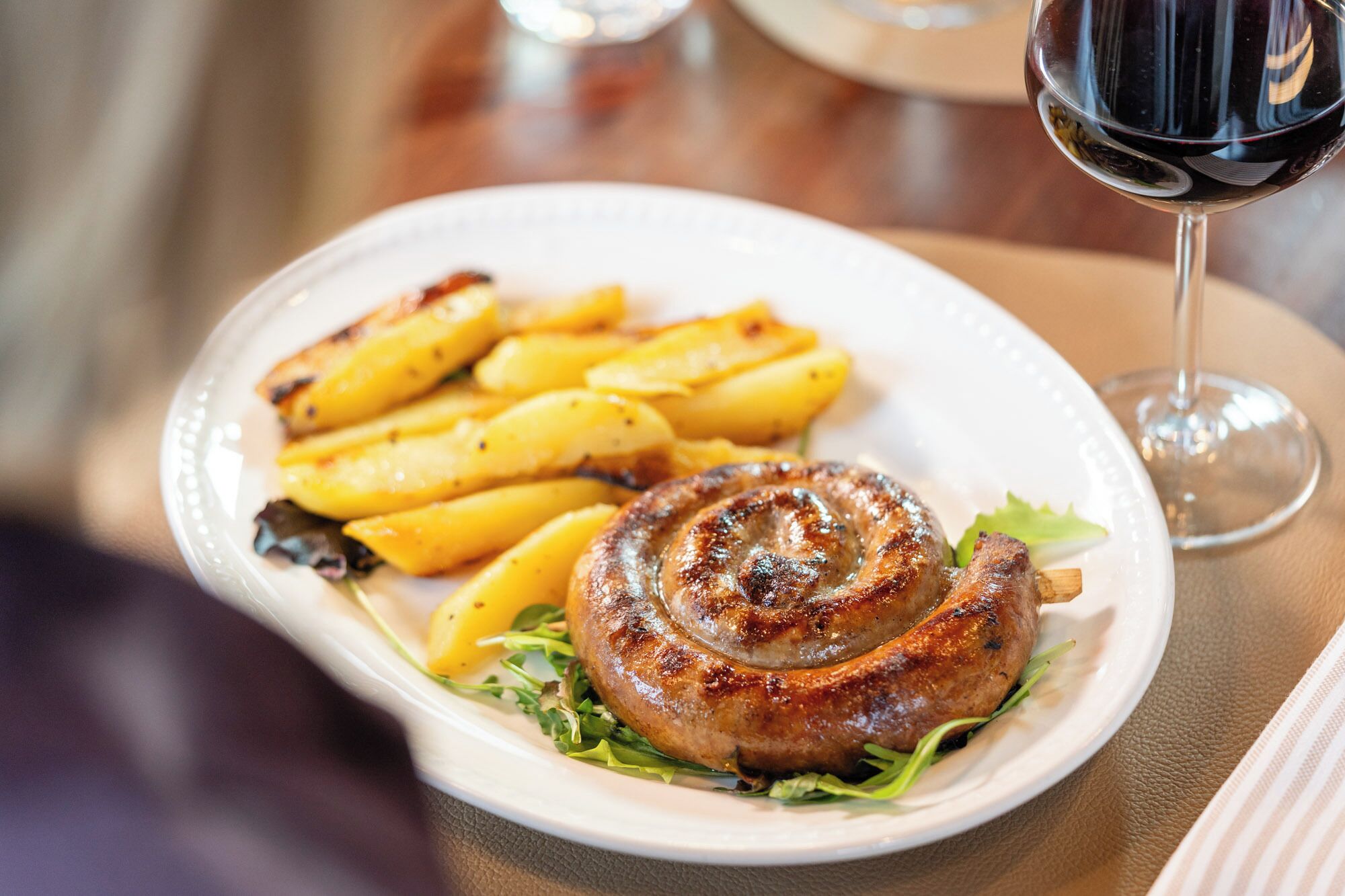 A plate with a grilled sausage and roasted potatoes, accompanied by a glass of red wine.