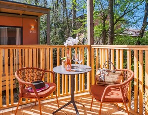 Una terraza de madera con dos sillas de mimbre, una mesita redonda con una botella de vino rosado y dos copas, y un bolso tejido.