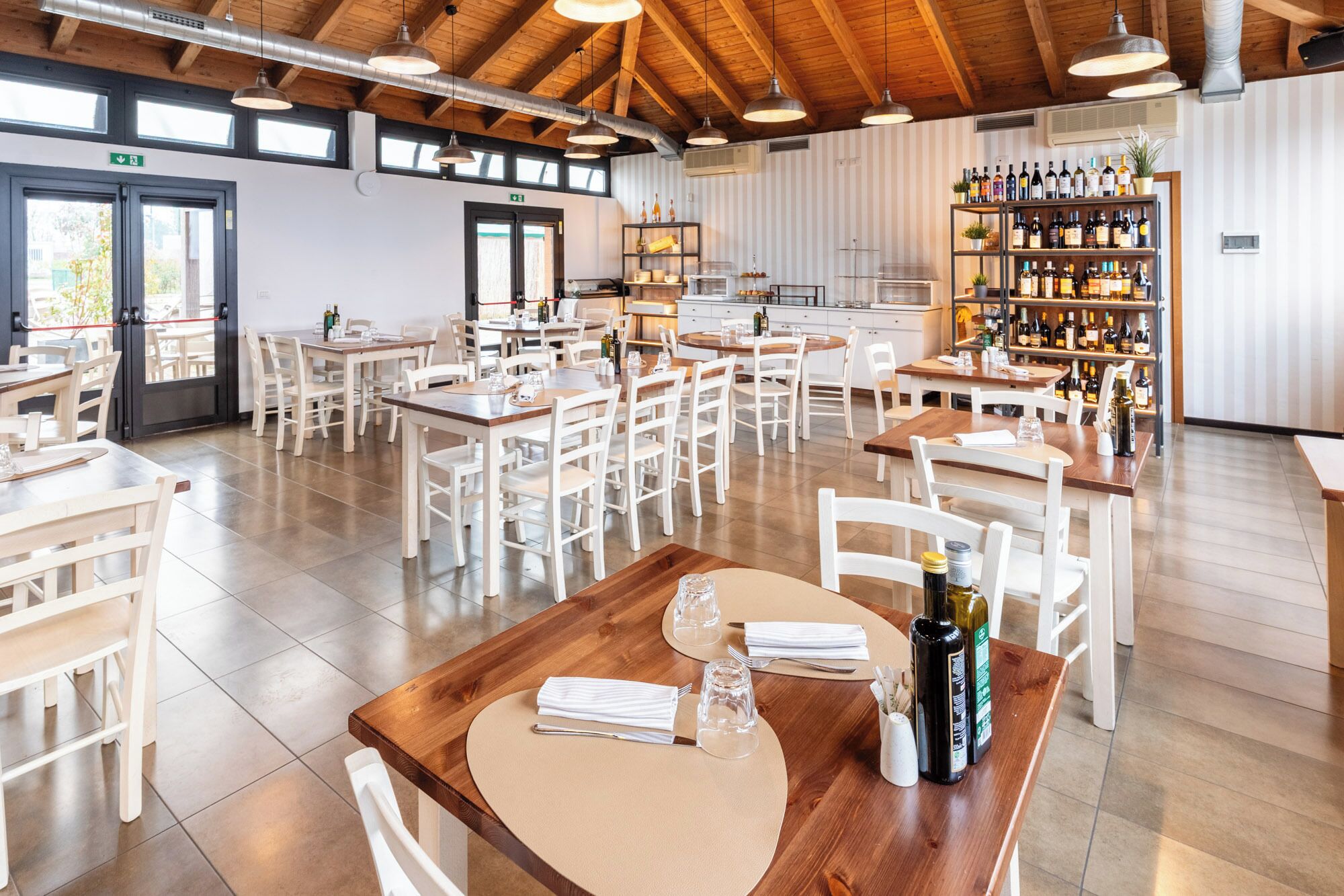 Un restaurante espacioso con numerosas mesas y sillas, una alacena para vinos y vistas al exterior a través de amplias ventanas.