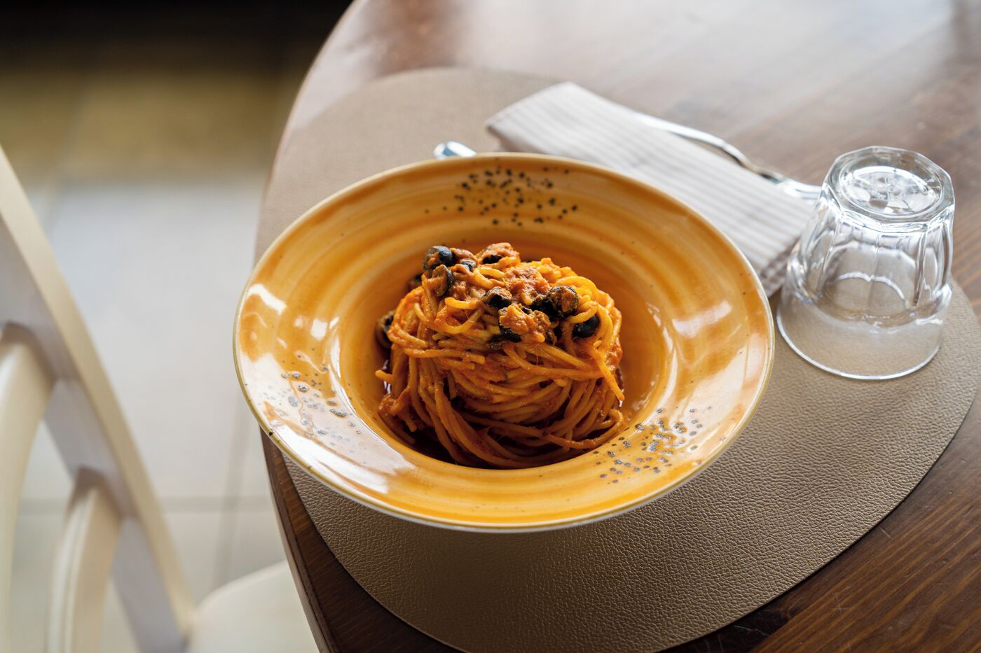A bowl of spaghetti with a tomato-based sauce, a clear glass, and a napkin on a table.