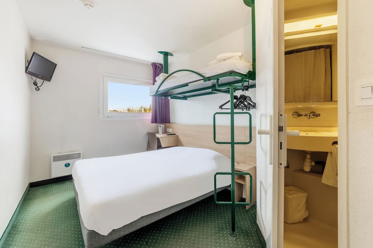 A compact hotel room with a bunk bed, a single bed, a small bedside table, a TV, a window with a view, and a bathroom with a shower and sink.