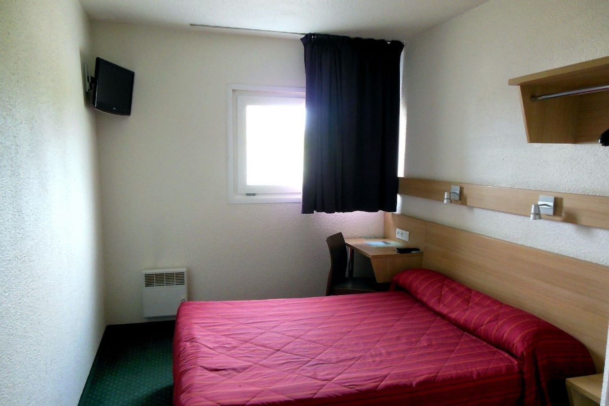 A small, single-bed room with a desk, chair, and a window with a black curtain.