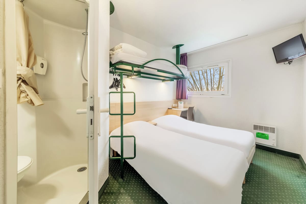 A hotel room with a bunk bed, a shower, a small table with a chair, a TV mounted on the wall, and a window with a view of trees.