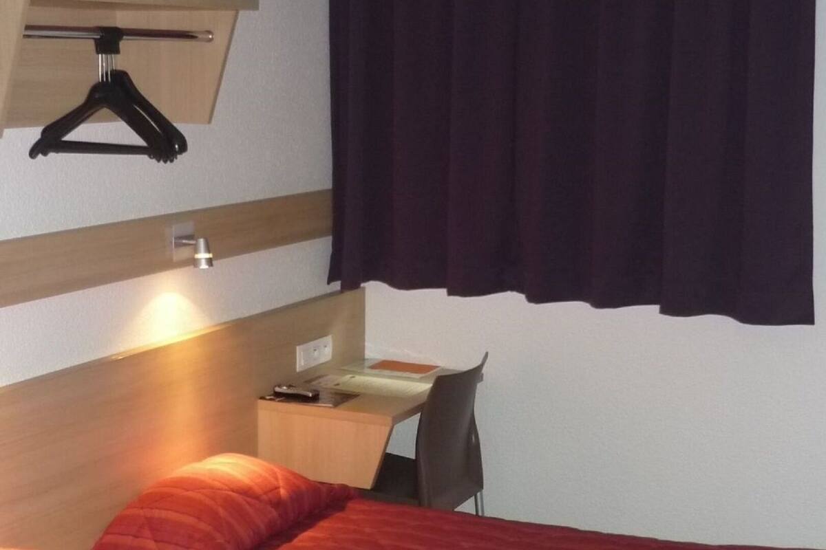 A hotel room with a bed, a desk, a chair, a hanging coat rack, and a window with curtains.