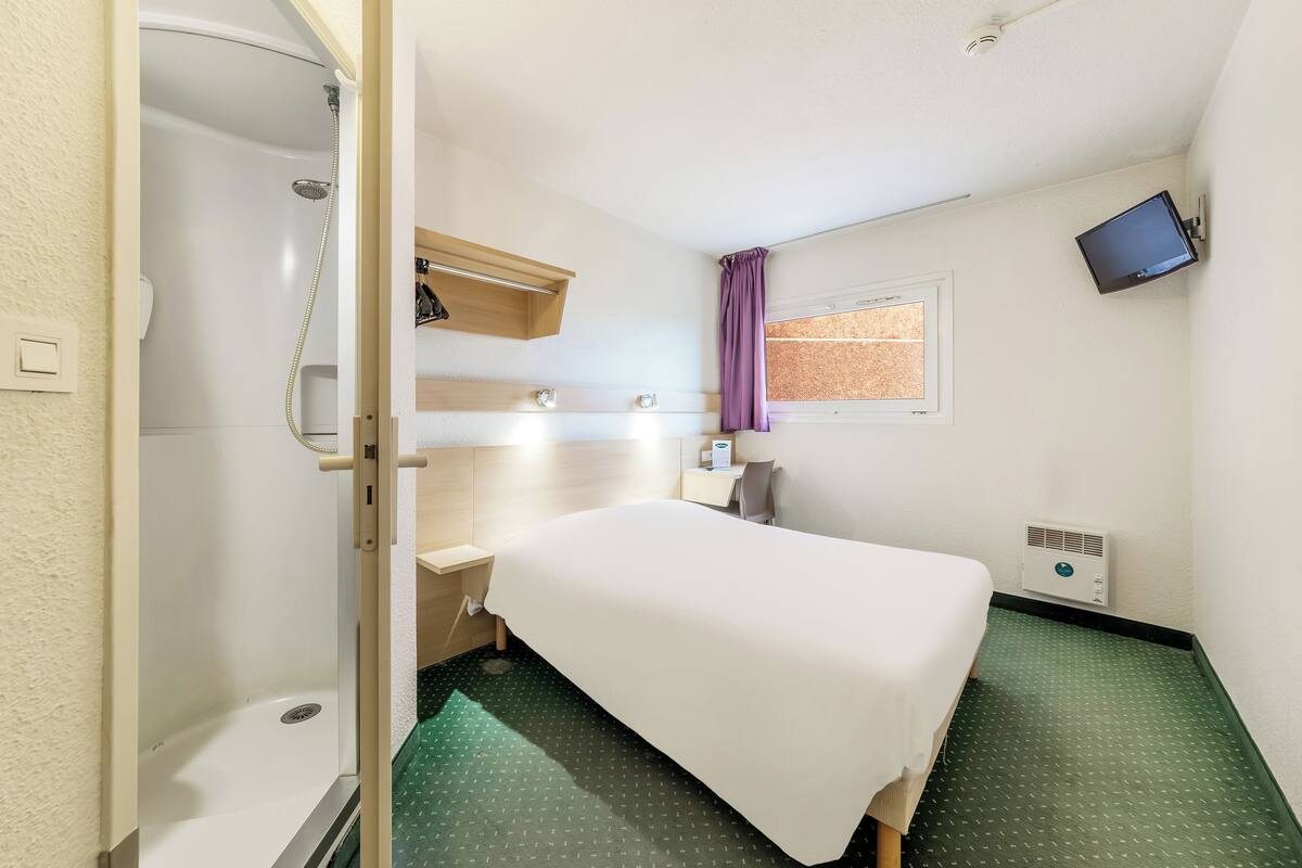 A hotel room with a bed, a shower, a small desk, a TV mounted on the wall, and a window with a curtain.