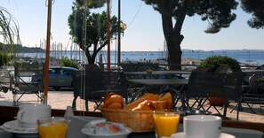 Un área de comedor al aire libre con mesas y sillas, con vista a un puerto deportivo con barcos y una playa al fondo.