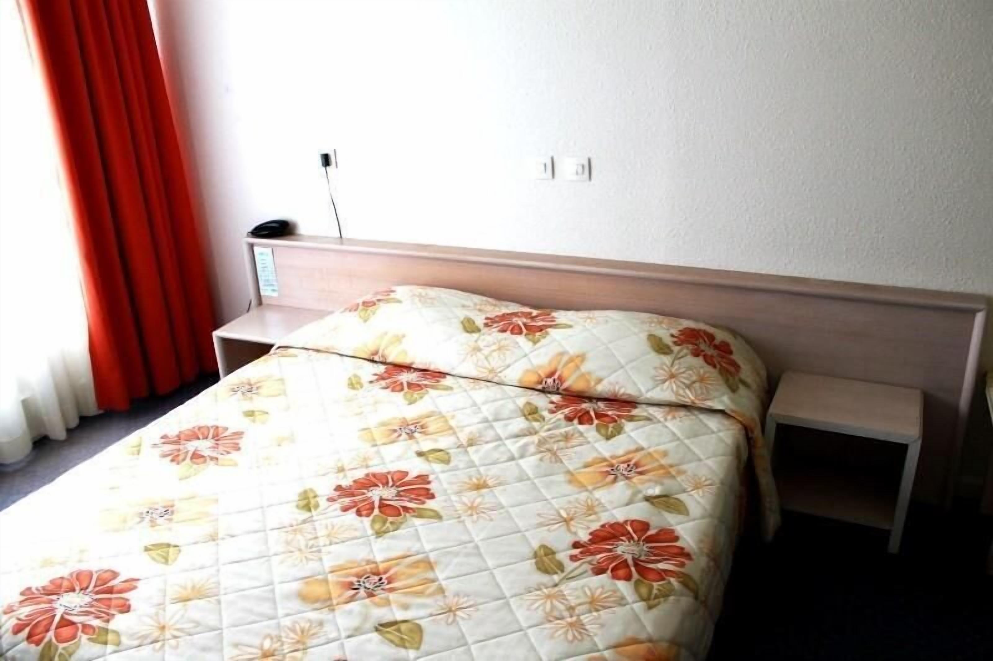 A neatly made bed with a floral duvet cover, a bedside table, and a power outlet on the wall.