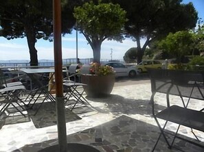 Un área de estar al aire libre con mesas y sillas, una planta grande en maceta y vista al mar.