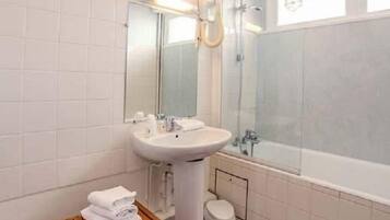 Un baño moderno con un lavamanos blanco, espejo y bañera. Hay un banco de madera con toallas y un cubo de basura blanco.