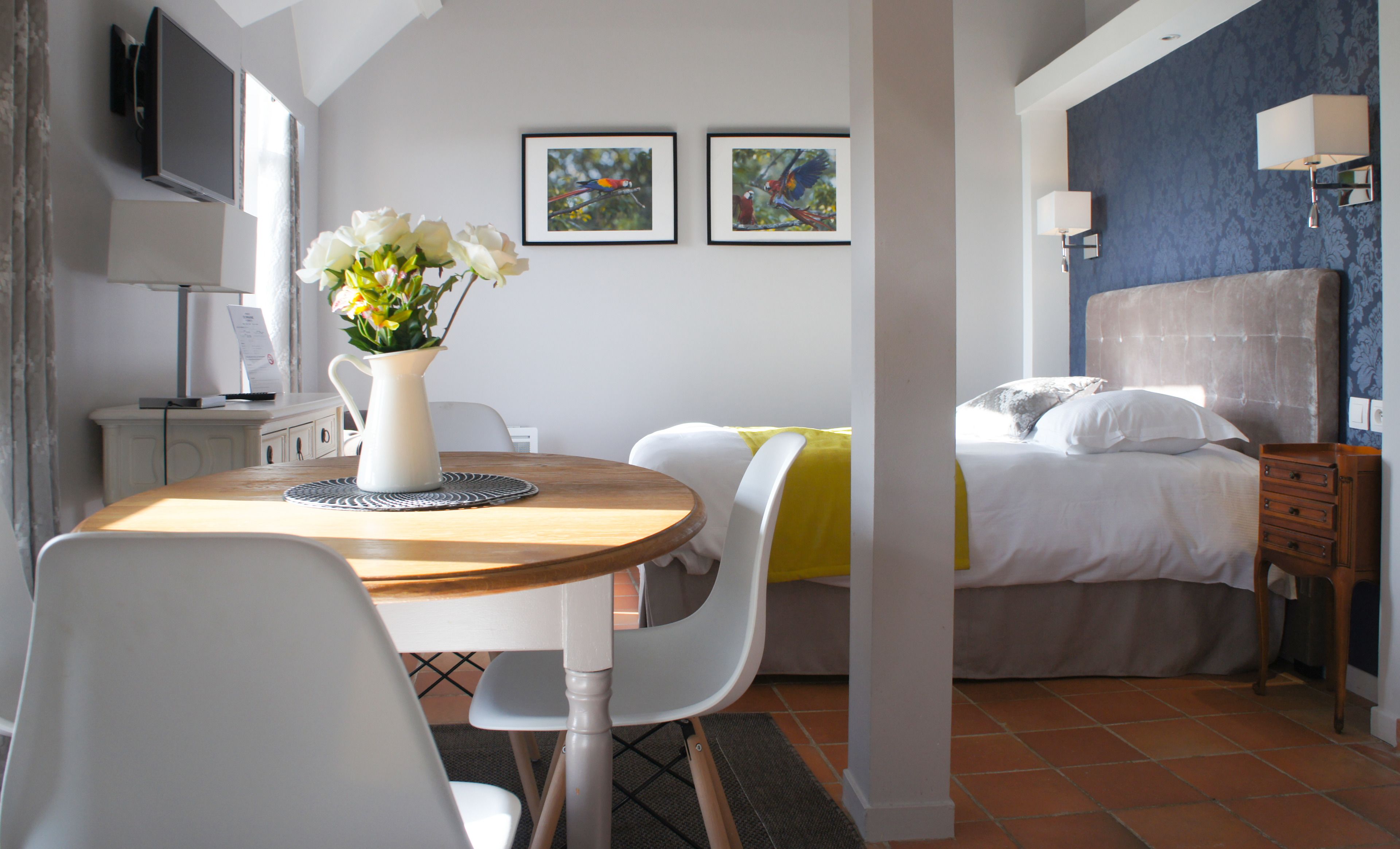 A hotel room with a bed, a desk with a computer, a chair, a vase with flowers, and two framed pictures on the wall.