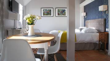 A hotel room with a bed, a desk with a computer, a chair, a vase with flowers, and two framed pictures on the wall.