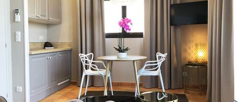 Una sala de estar moderna con una mesa redonda, dos sillas blancas, un sofá, un televisor y una ventana con cortinas.