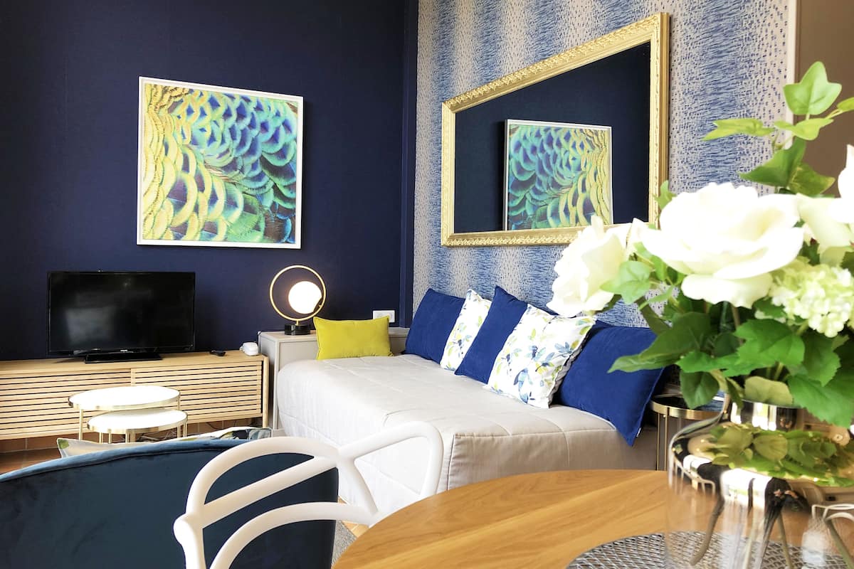 Una habitación de hotel con una cama, televisión, mesita de noche con lámpara, espejo y un jarrón con flores.