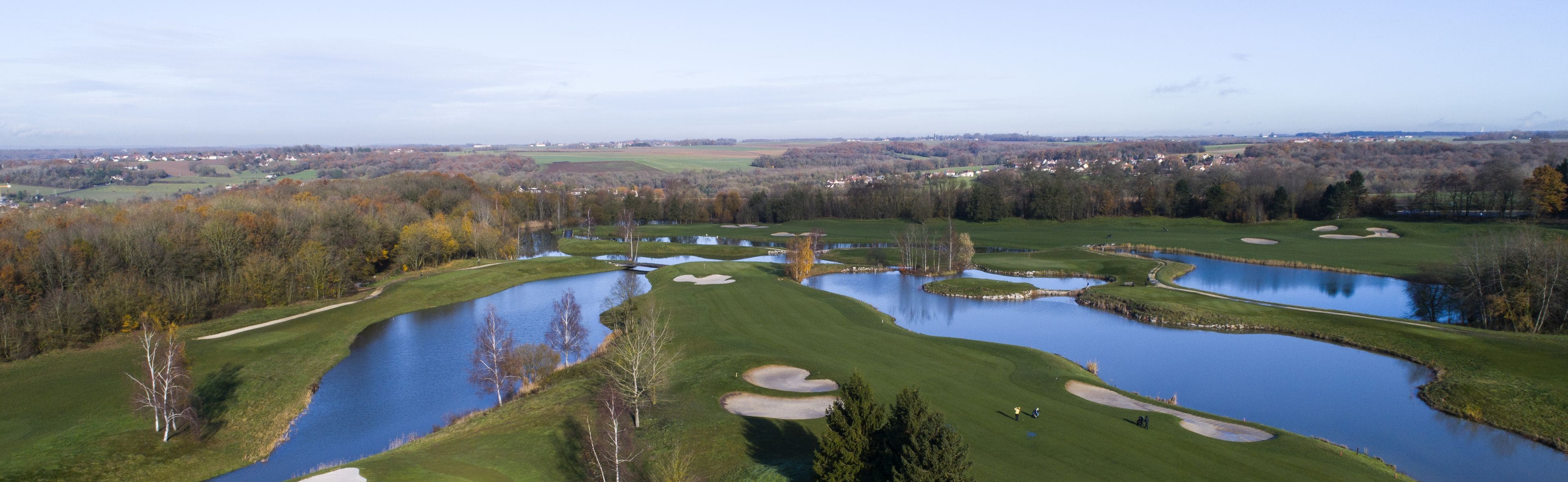 A golf course with multiple ponds and sand bunkers, surrounded by trees and a distant town.