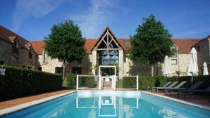 A swimming pool with a stone building in the background, a covered area with umbrellas, and a clear sky. - Hotel Les Suites - De Crécy (Crecy-la-Chapelle)