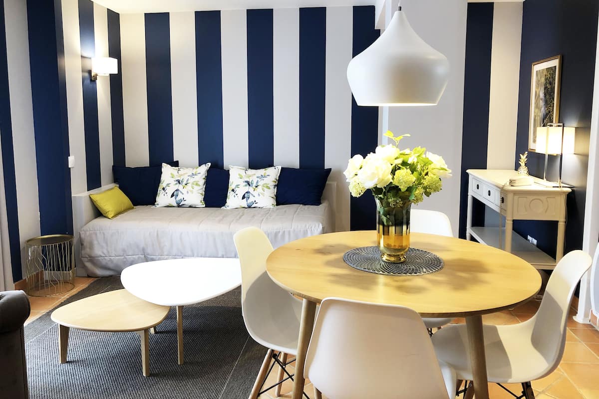 Una habitación de hotel moderna con una cama, una mesa de comedor con sillas, un escritorio pequeño y un florero con flores.