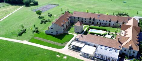 Vista aérea de una extensa propiedad con varias edificaciones, incluyendo una casa principal con fachada de piedra, una piscina y céspedes bien cuidados.
