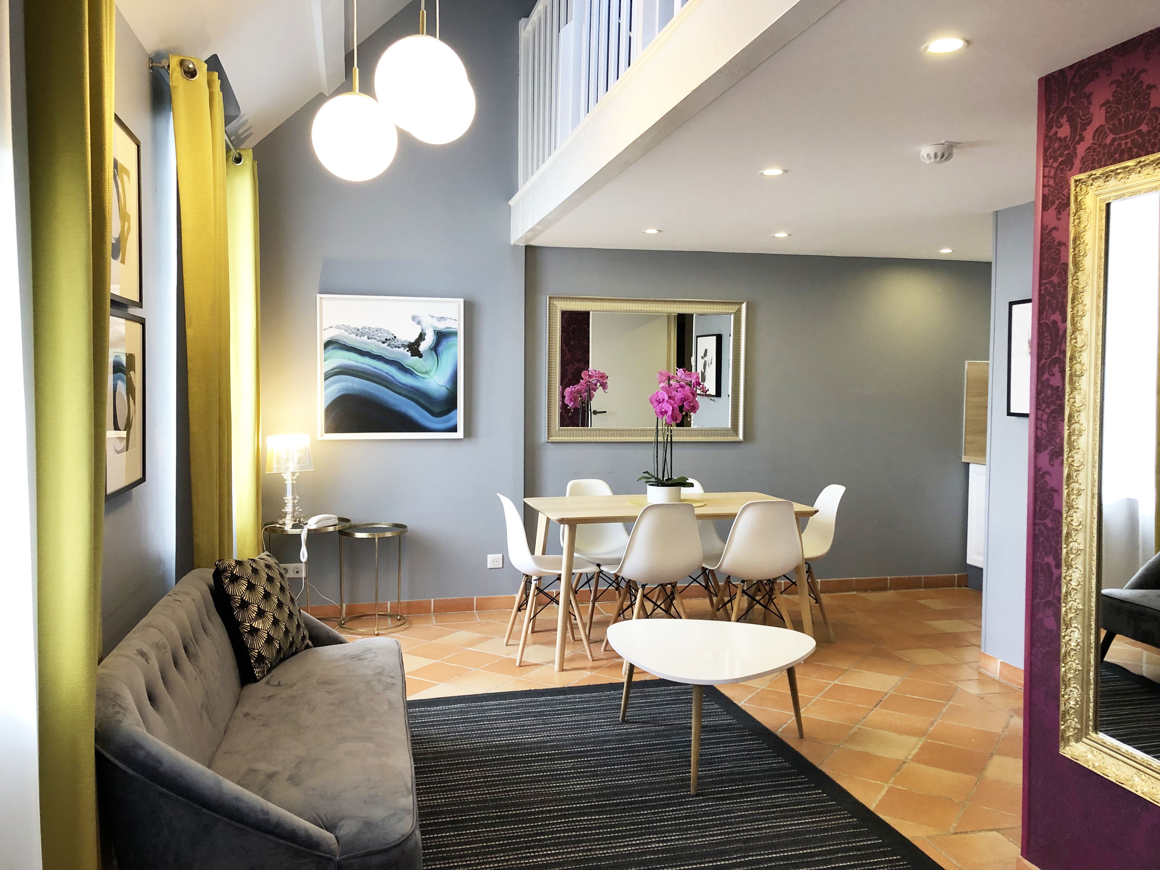 A modern living room with a grey sofa, a dining table with chairs, a black rug, and a large mirror on the wall.