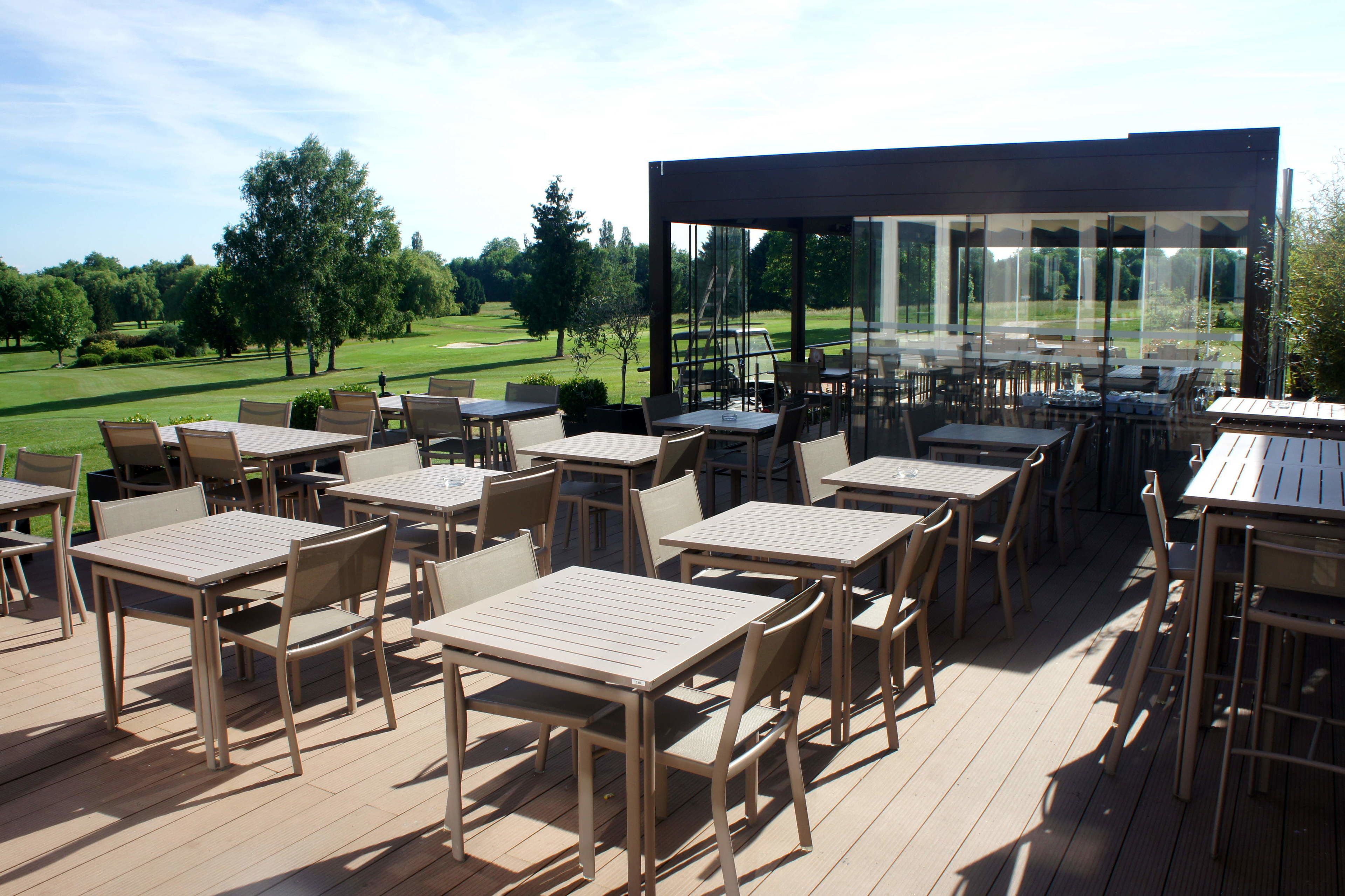 An outdoor patio with tables and chairs, a glass enclosure, and a view of a golf course.