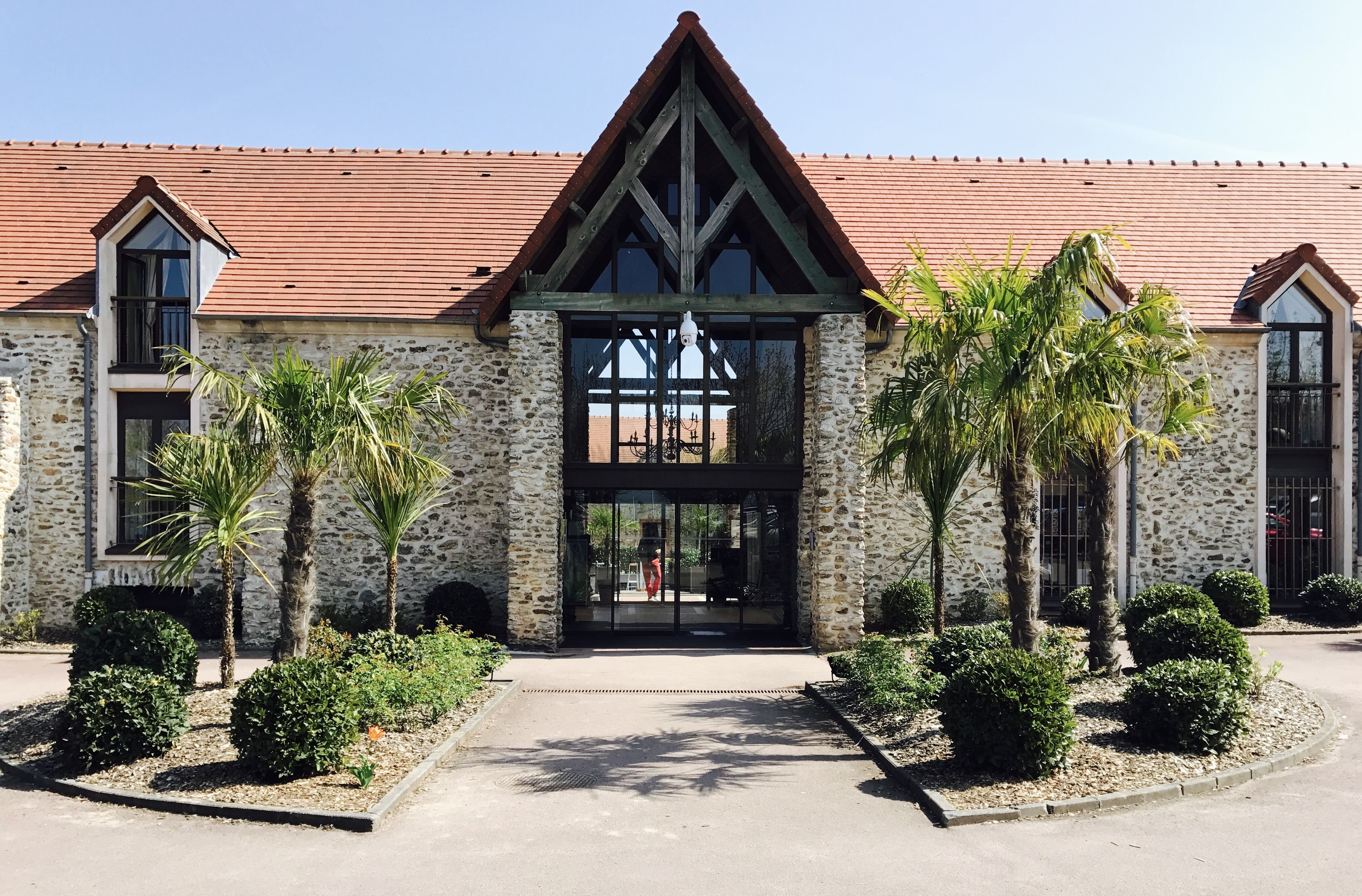 A stone building with a red-tiled roof, a black entrance, and palm trees in the foreground.