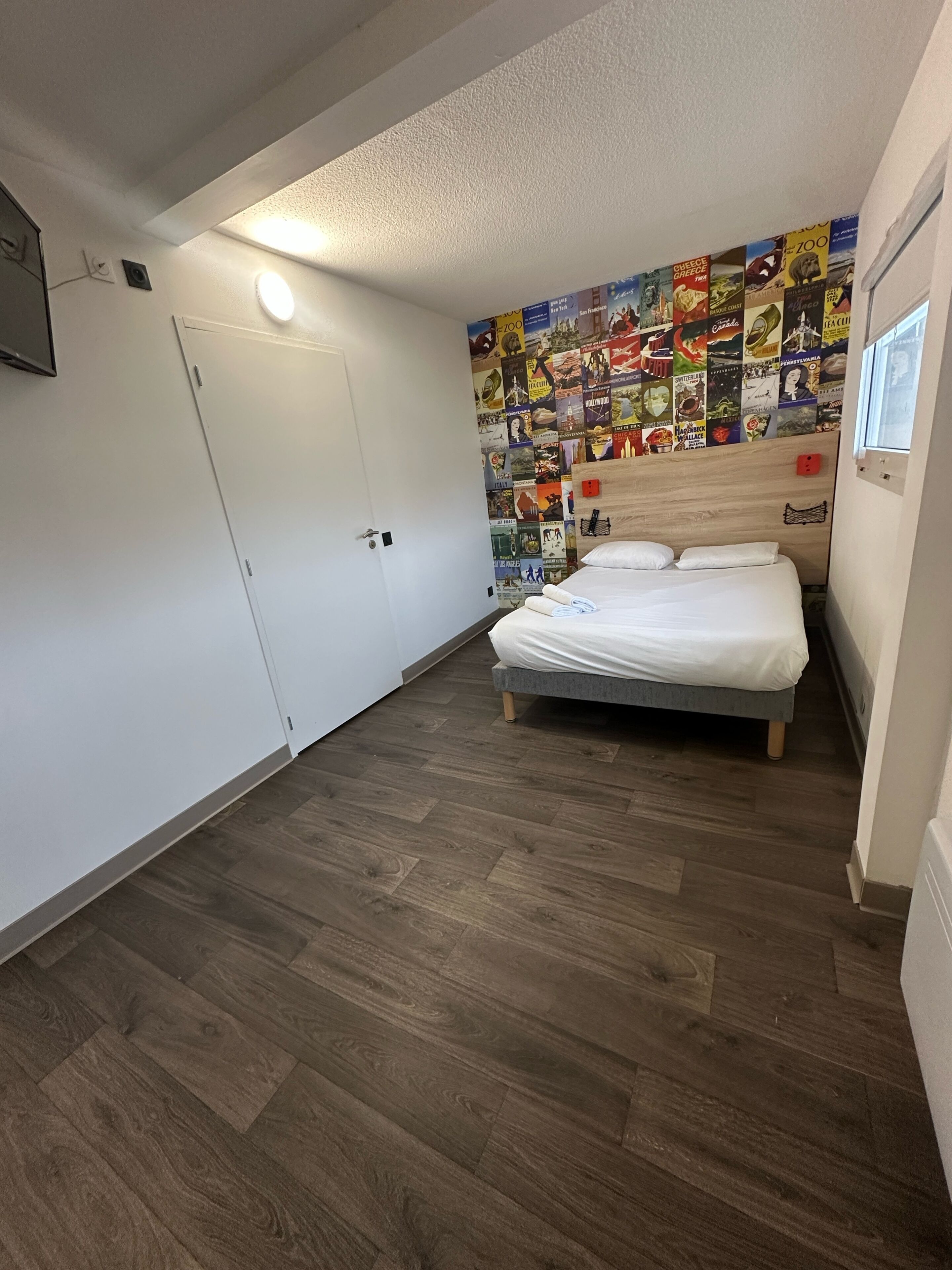 Un dormitorio con cama, suelo de madera, una pared con carteles de viajes y una puerta.