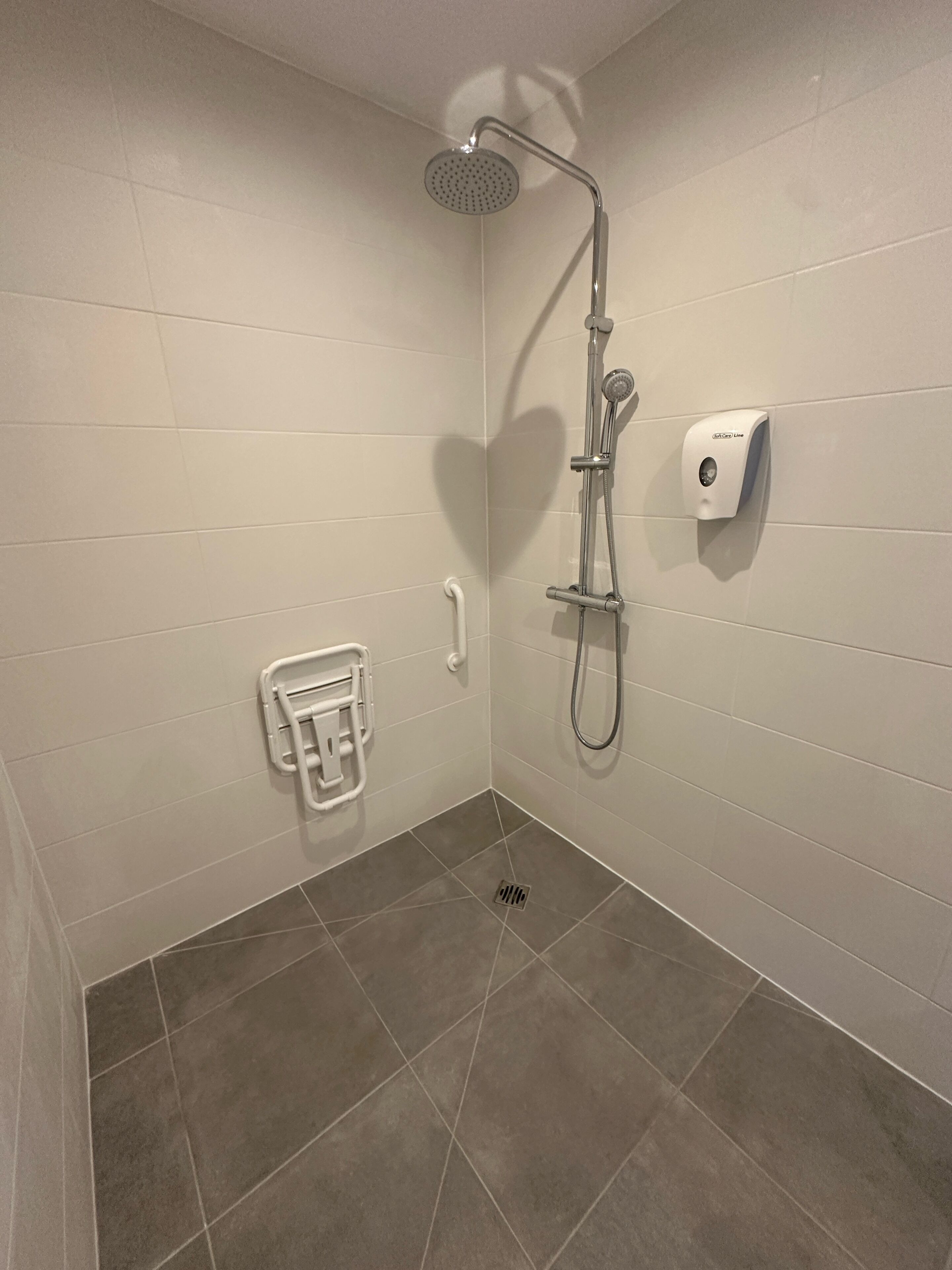Un baño con paredes de azulejos blancos, ducha con cabezal de ducha manual, toallero y dispensador de jabón.