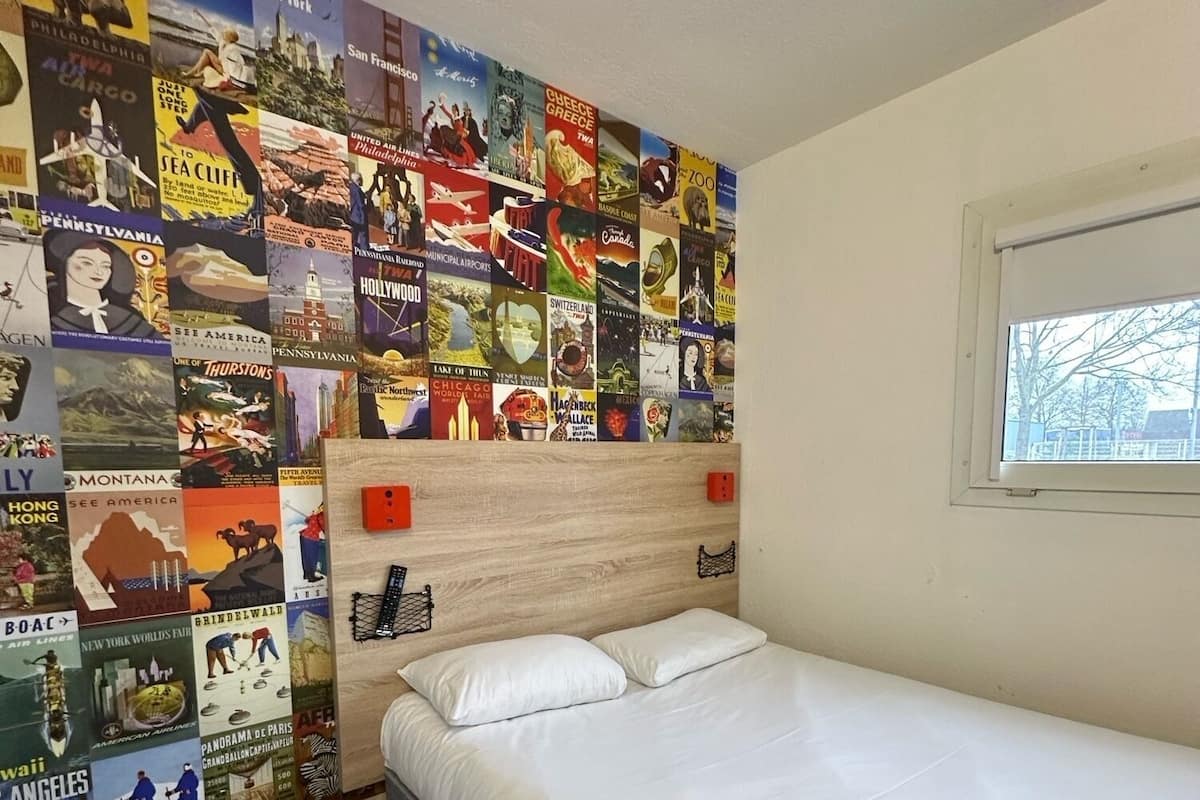 A hotel room with a bed, pillows, a headboard with a poster collage, a window, and a wooden floor.