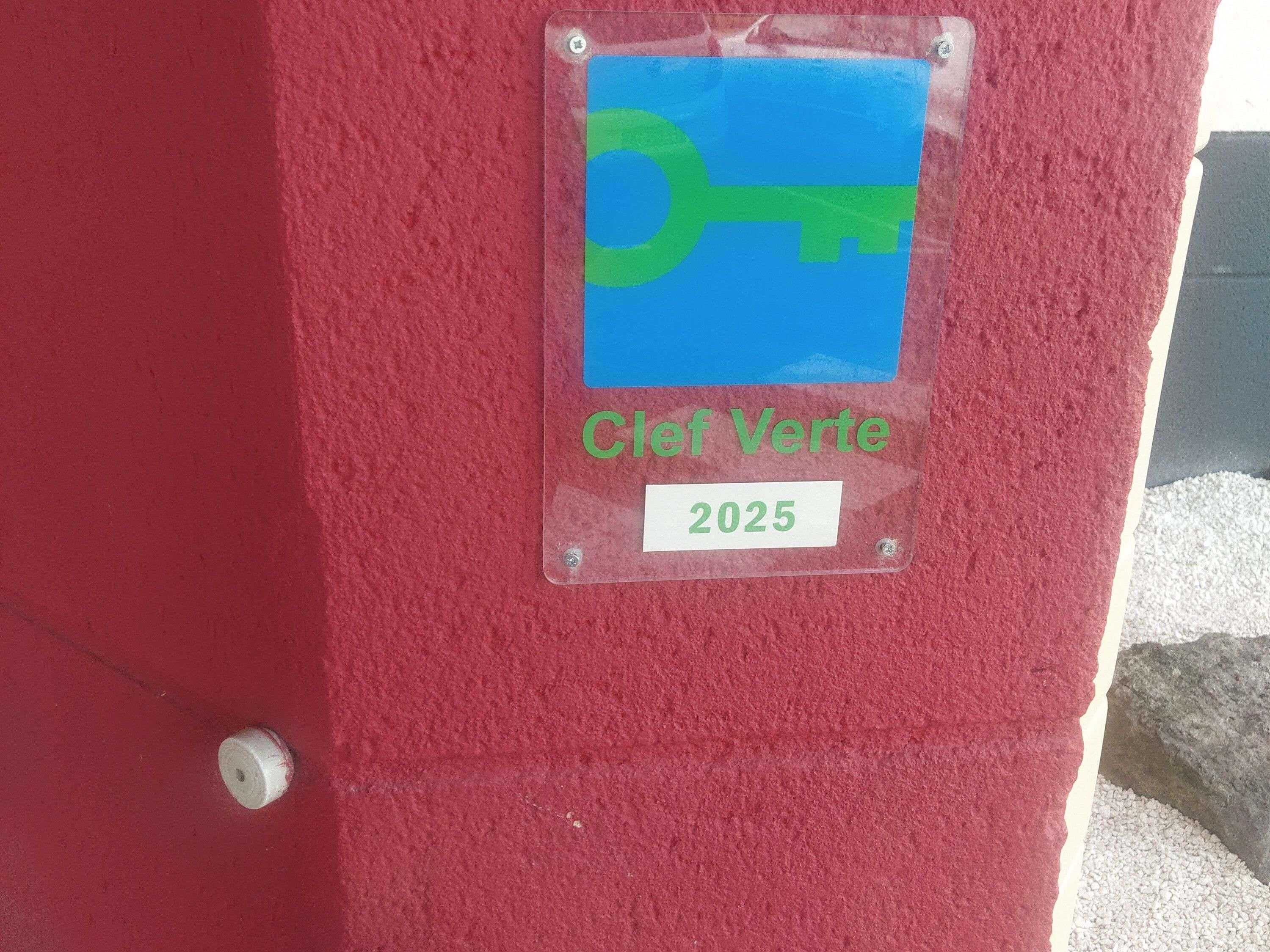 Un mur rouge sur lequel est fixé un panneau transparent en plastique, orné d’un symbole de clé verte et du texte « Cif Verde 2025 ».