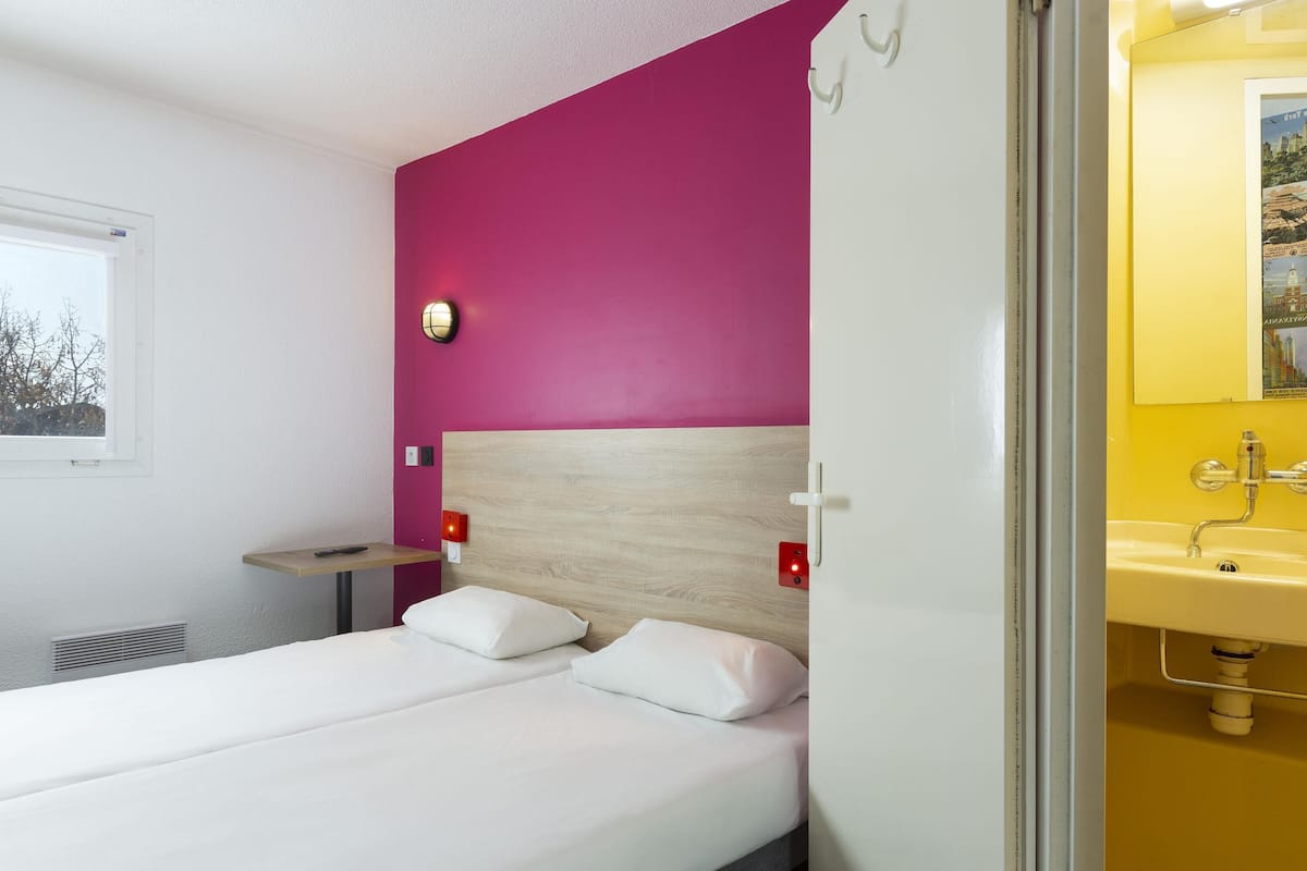 A hotel room with a bed, a nightstand, a wall-mounted lamp, and a bathroom with a sink and mirror.