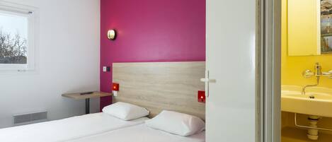 Habitación de hotel con cama, mesita de noche, lámpara de pared y baño con lavabo y espejo.