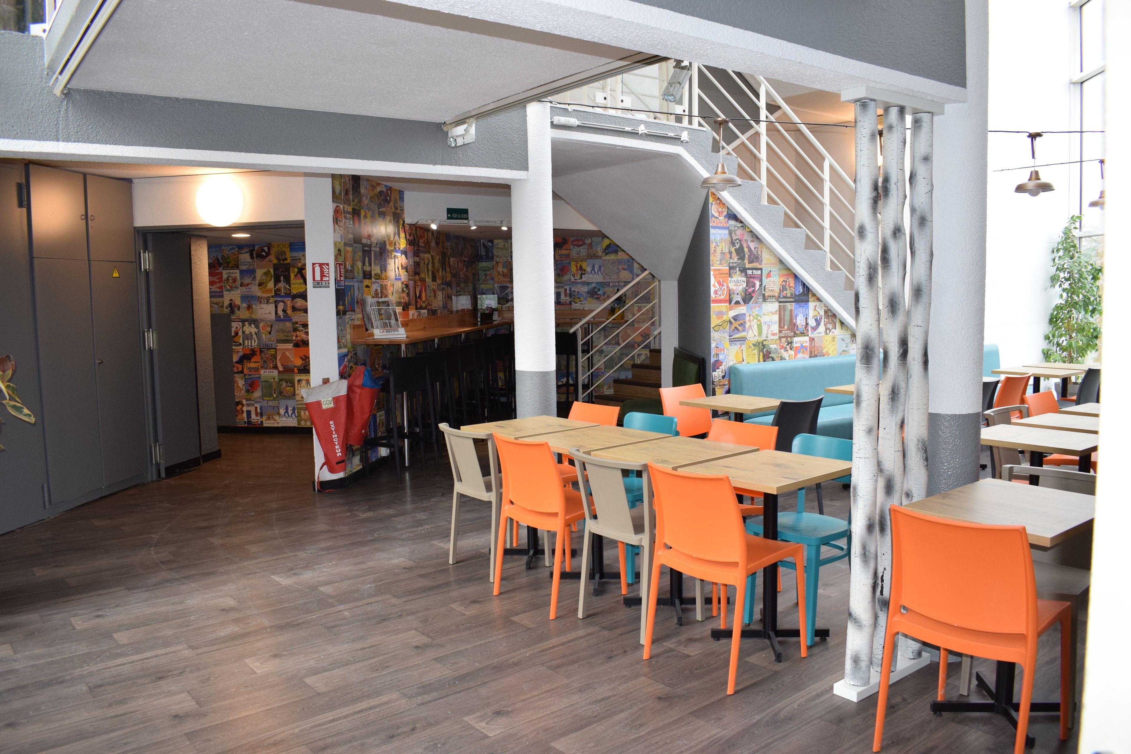 A modern restaurant interior with wooden flooring, orange and blue chairs, a staircase, and a wall decorated with colorful posters.