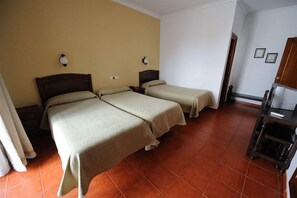 Habitación de hotel con dos camas individuales, una mesita de noche de madera con lámpara, una pequeña mesa con una silla y un armario.