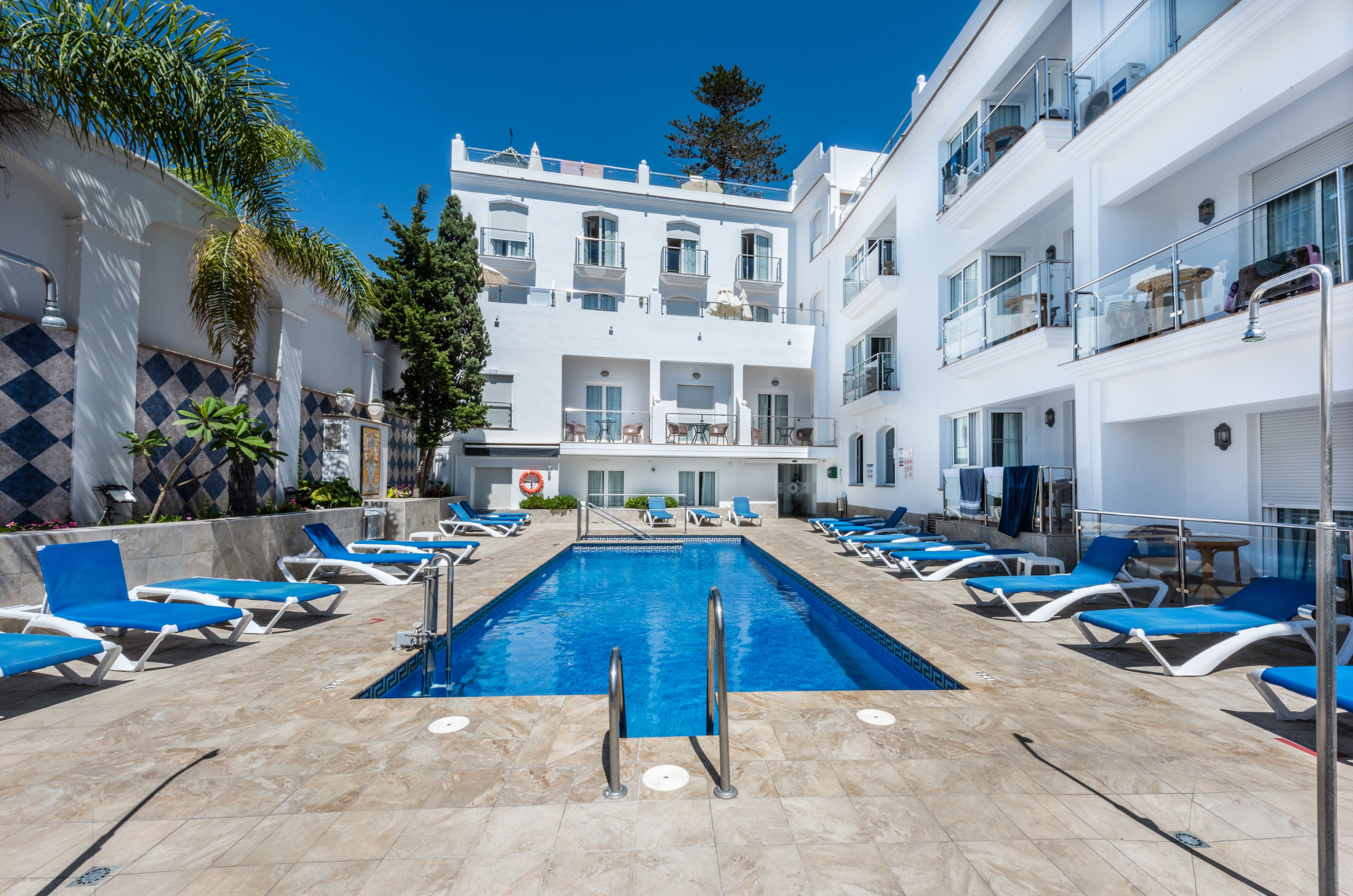A pool area with blue lounge chairs, a clear blue pool, and a white building with balconies.