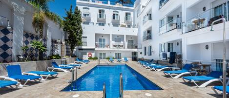 Un área de piscina con sillas reclinables azules, una piscina azul cristalina y un edificio blanco con balcones.