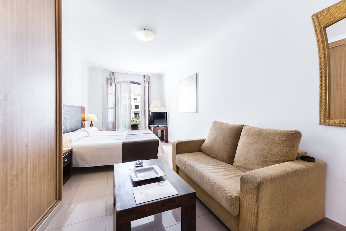 A hotel room with a bed, a sofa, a table, a TV, and a window with curtains.