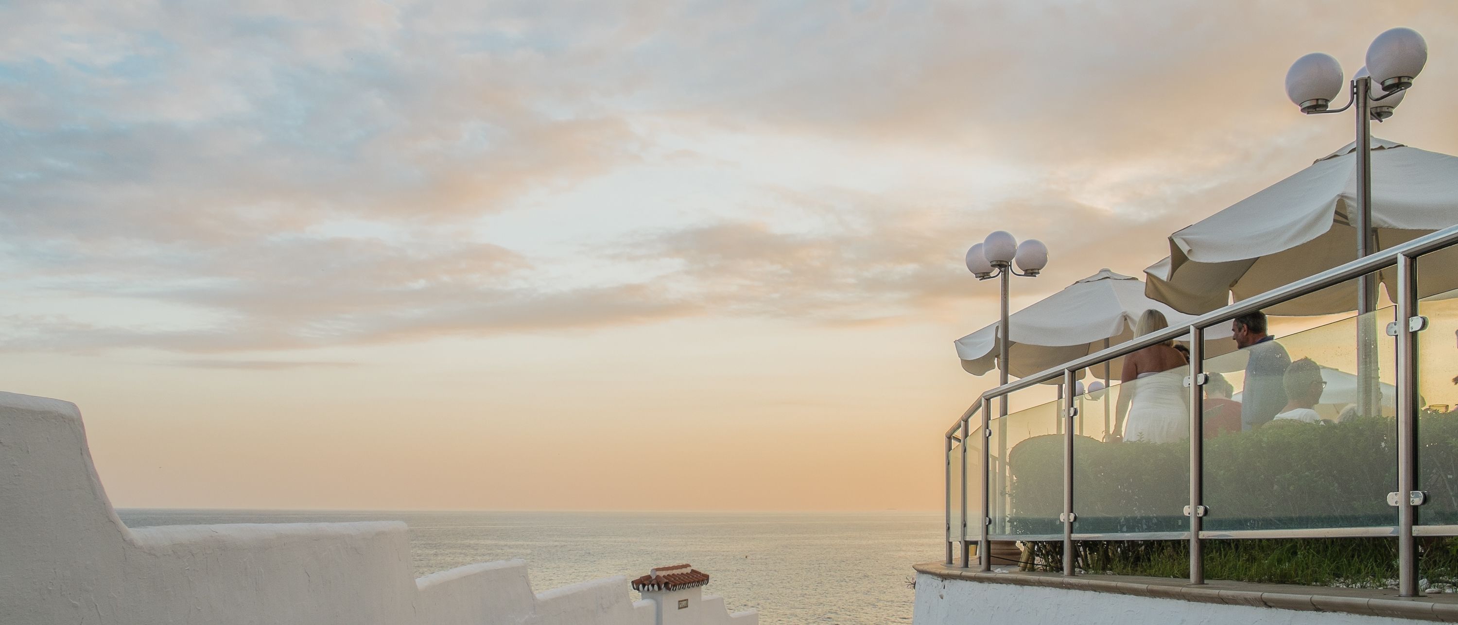 A seaside terrace with a glass balustrade, white umbrellas, and a clear view of the ocean.