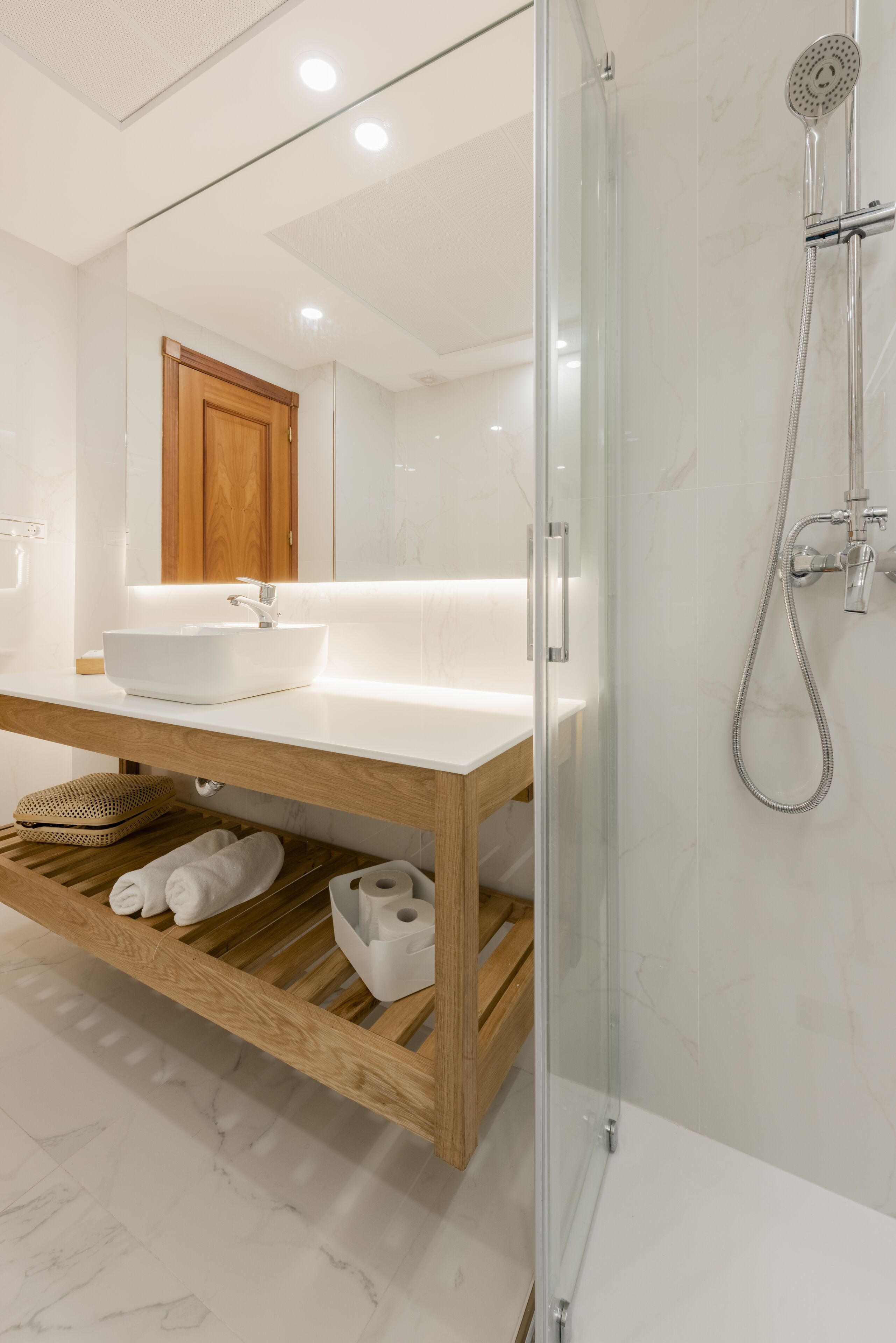 Un baño moderno con cabina de ducha de vidrio, tocador de madera con lavamanos blanco y suelo de mármol.
