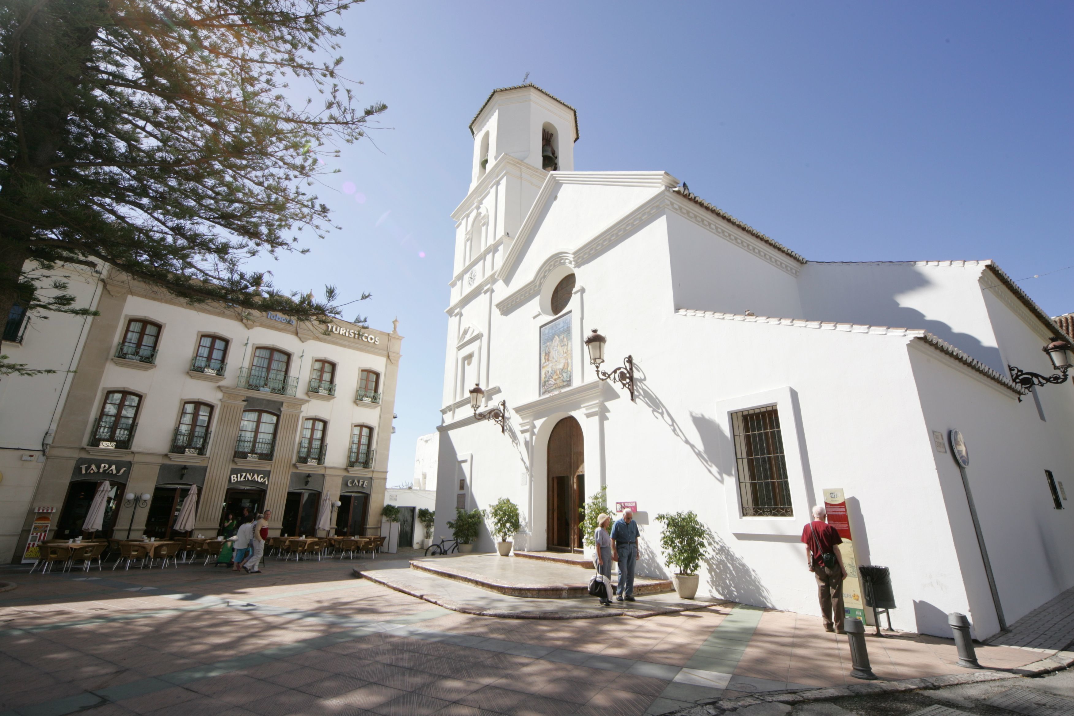 A plaza with a white building featuring a bell tower, surrounded by cafes and outdoor seating.