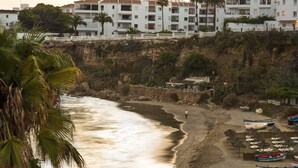 Una playa con barcos, una persona caminando y edificios al fondo.