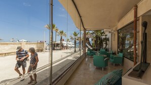 Un hotel frente a la playa con pared de vidrio que da al mar, asientos al aire libre y palmeras.