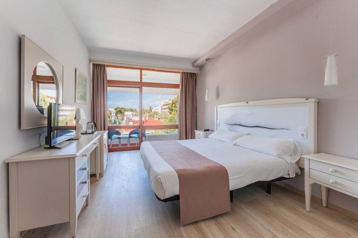 Een hotelkamer met een bed, een bureau met een televisie, een stoel en uitzicht op gebouwen door grote ramen.
